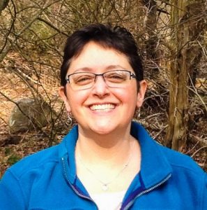 A person with short dark hair and glasses smiles outdoors, wearing a blue jacket and a necklace, with trees and branches in the background.