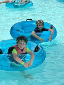 Two smiling children wearing swim goggles relax on blue inflatable tubes in a pool. Both boys have wristbands and short-sleeve swim shirts. The clear water sparkles around them on a sunny day.