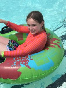 A smiling girl wearing a pink and orange striped long-sleeve swim shirt floats on a colorful inflatable ring in a swimming pool.