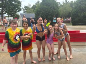 Six children in swimsuits smile and pose together at an outdoor water park, standing on wet pavement with trees and water attractions visible in the background. Some are wearing colorful shirts, and everyone looks happy.
