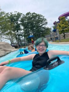 A smiling child with short blue-green hair floats in a blue inner tube on a lazy river at a water park, with trees and water slides visible in the background. Another child floats behind them.