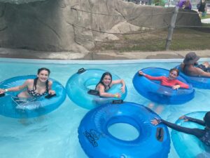 Three girls smile and float on blue inner tubes in a lazy river at a water park, with other people nearby and a large rock structure in the background.