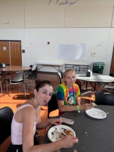 Two teenage girls sit at a table eating from paper plates in a cafeteria. One wears a white tank top, the other a colorful tie-dye shirt. They are looking at the camera, and there are empty tables and a whiteboard in the background.