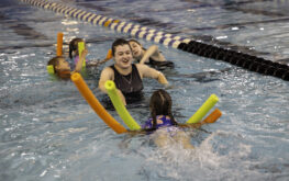 A swim instructor in a pool smiles while teaching several children who are using colorful pool noodles to practice swimming. The group is surrounded by lane dividers in an indoor swimming pool.