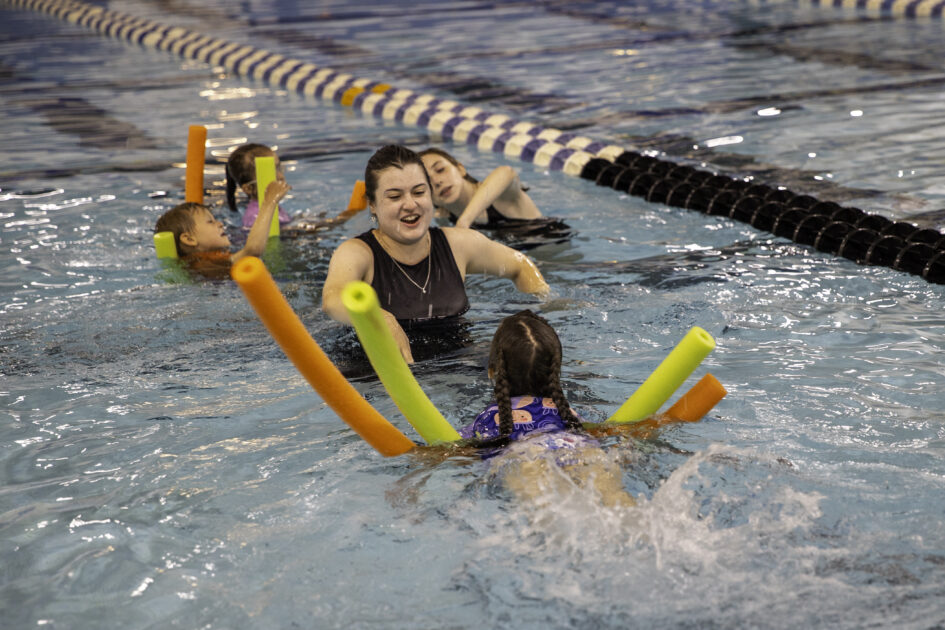 A swim instructor in a pool smiles while teaching several children who are using colorful pool noodles to practice swimming. The group is surrounded by lane dividers in an indoor swimming pool.