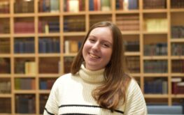 A young woman with long brown hair, wearing a white sweater with black stripes, smiles in front of a wooden bookshelf filled with books.