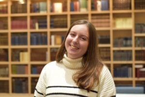 A young woman with long brown hair, wearing a white sweater with black stripes, smiles in front of a wooden bookshelf filled with books.